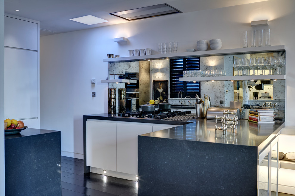 Small contemporary l-shaped kitchen/diner in London with flat-panel cabinets, white cabinets, marble worktops, multi-coloured splashback, mirror splashback, black appliances, dark hardwood flooring and a breakfast bar.