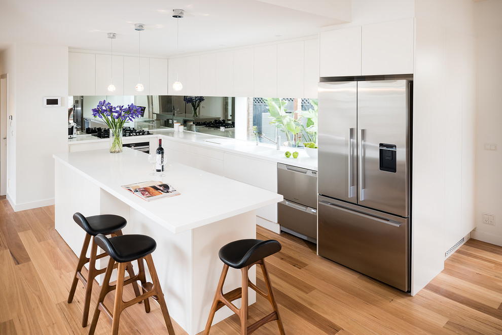 Mid-sized trendy l-shaped medium tone wood floor eat-in kitchen photo in Melbourne with an integrated sink, flat-panel cabinets, white cabinets, solid surface countertops, mirror backsplash, stainless steel appliances, an island, metallic backsplash and white countertops
