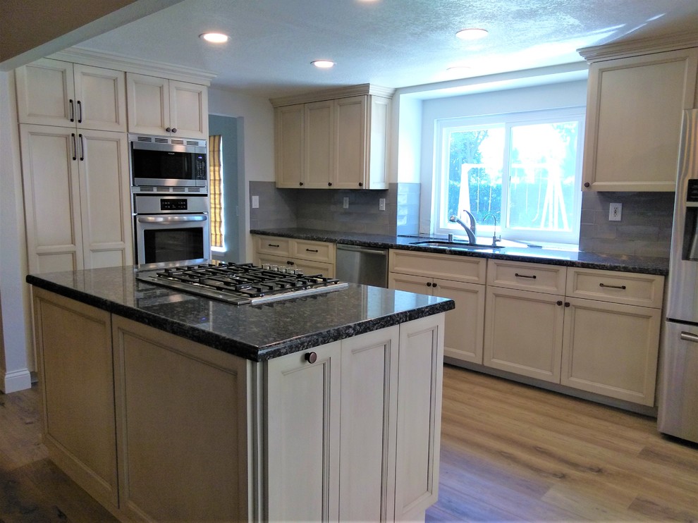 Example of a mid-sized transitional galley vinyl floor and brown floor open concept kitchen design in Los Angeles with an undermount sink, recessed-panel cabinets, beige cabinets, granite countertops, gray backsplash, ceramic backsplash, stainless steel appliances, an island and black countertops