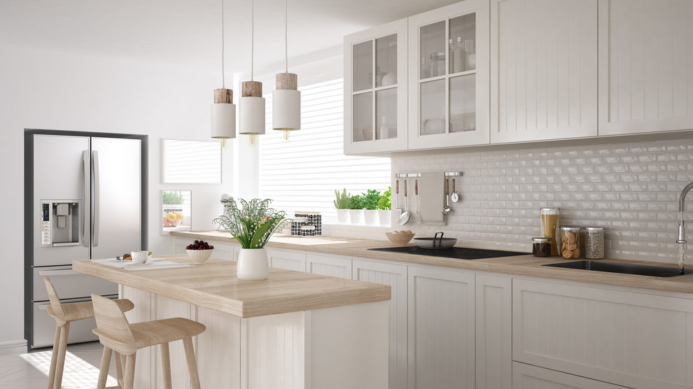 Example of a mid-sized cottage chic single-wall light wood floor and beige floor enclosed kitchen design in Miami with a drop-in sink, louvered cabinets, white cabinets, wood countertops, white backsplash, porcelain backsplash, stainless steel appliances and an island