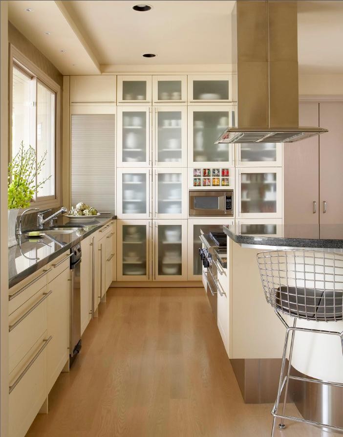 Modern inredning av ett kök, med en undermonterad diskho, luckor med glaspanel, vita skåp, granitbänkskiva, rostfria vitvaror, ljust trägolv och en köksö