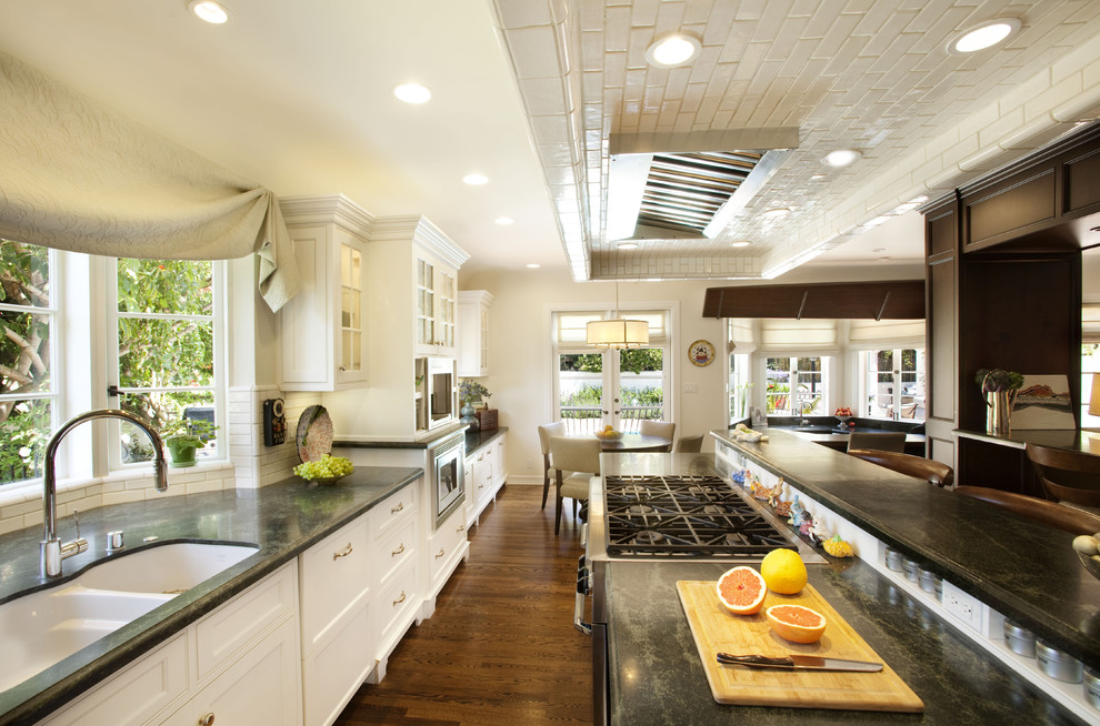 This is an example of a large beach style galley open plan kitchen in Los Angeles with a submerged sink, recessed-panel cabinets, white cabinets, white splashback, metro tiled splashback, stainless steel appliances, medium hardwood flooring, an island, soapstone worktops and brown floors.