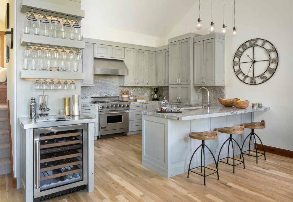 Bild på ett rustikt grå grått u-kök, med skåp i shakerstil, grå skåp, grått stänkskydd, stänkskydd i sten, rostfria vitvaror, mellanmörkt trägolv, en halv köksö och brunt golv