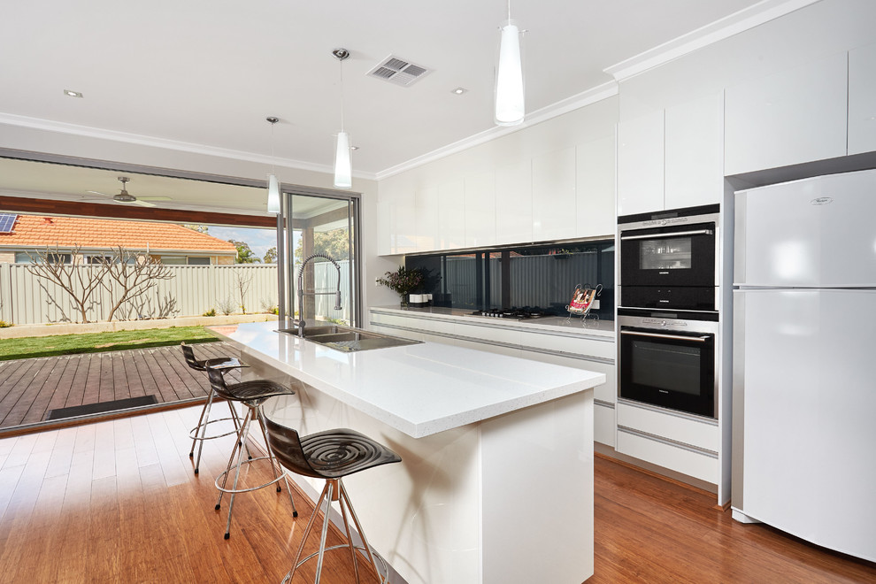 Large modern single-wall kitchen/diner in Perth with a double-bowl sink, flat-panel cabinets, white cabinets, glass sheet splashback, black appliances, bamboo flooring, an island, composite countertops, black splashback and white worktops.