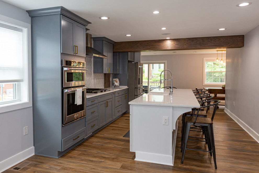 Photo of a large classic single-wall kitchen/diner in DC Metro with a belfast sink, blue cabinets, ceramic splashback, stainless steel appliances, light hardwood flooring, an island, brown floors and white worktops.