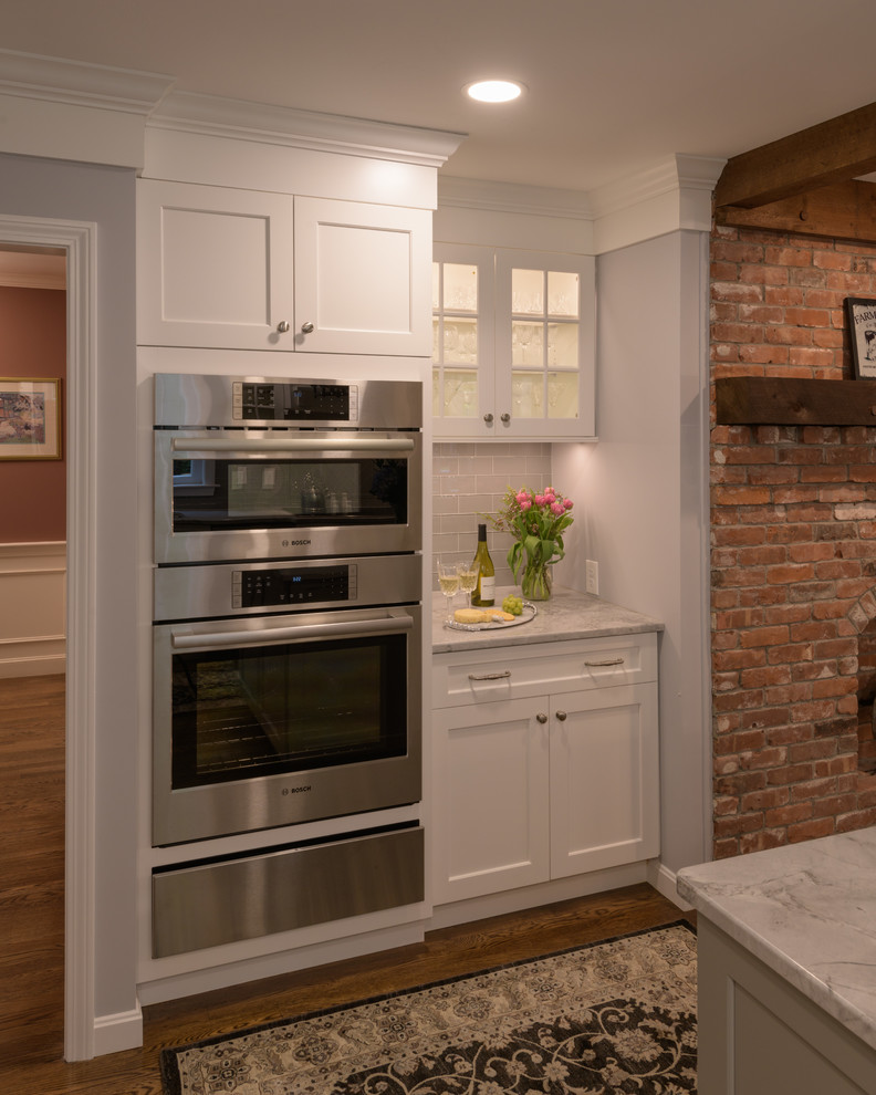 Inredning av ett rustikt mellanstort vit vitt kök, med en undermonterad diskho, luckor med infälld panel, vita skåp, marmorbänkskiva, grått stänkskydd, stänkskydd i tunnelbanekakel, rostfria vitvaror, mörkt trägolv, en köksö och brunt golv
