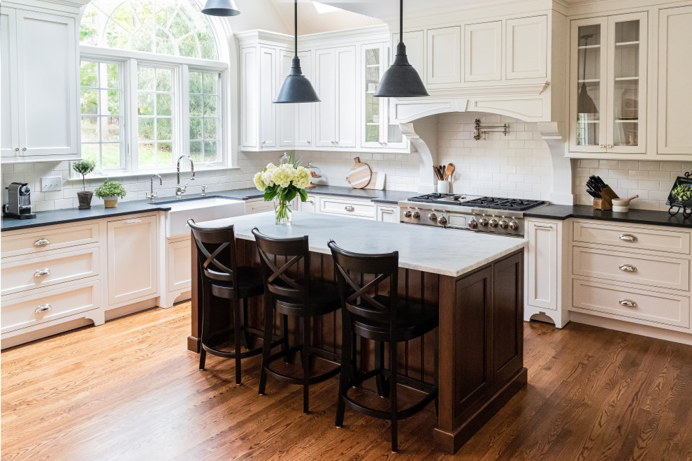 Large traditional u-shaped open plan kitchen in Philadelphia with a farmhouse sink, beaded inset cabinets, white cabinets, granite benchtops, white splashback, subway tile splashback, panelled appliances, medium hardwood floors, with island, brown floor and black benchtop.