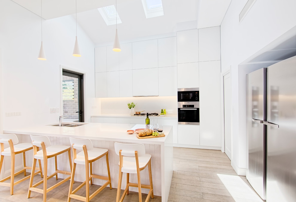 Große Moderne Wohnküche in L-Form mit Doppelwaschbecken, flächenbündigen Schrankfronten, weißen Schränken, Küchenrückwand in Weiß, Glasrückwand, Küchengeräten aus Edelstahl, hellem Holzboden und Kücheninsel in Sydney