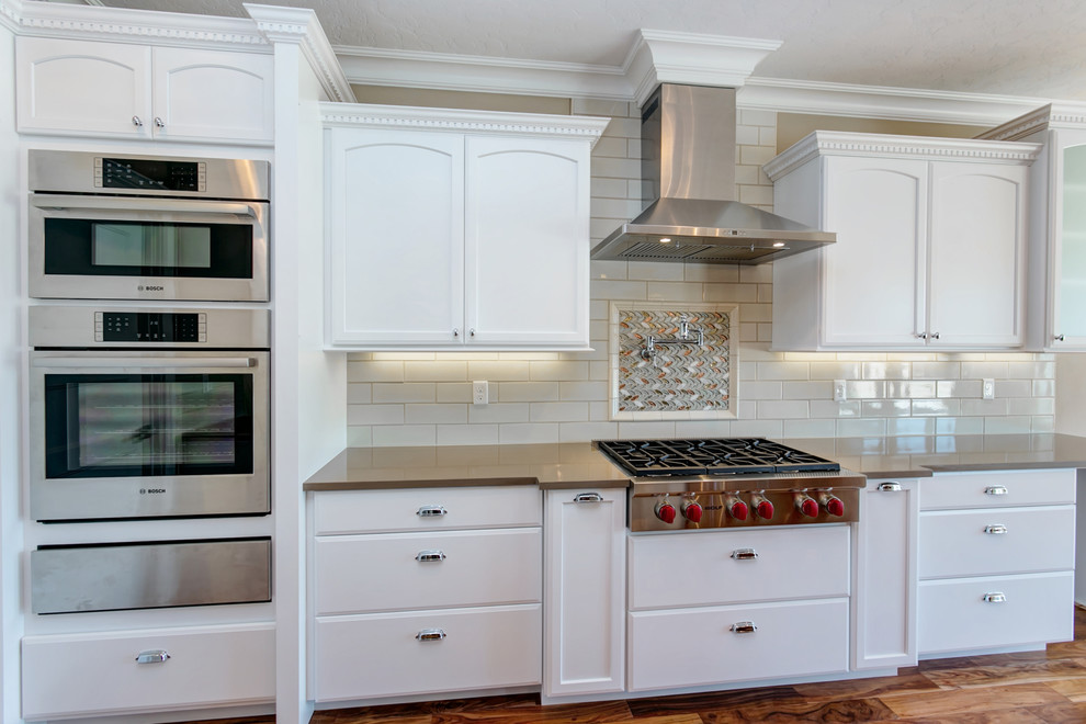 Exempel på ett stort klassiskt kök, med en undermonterad diskho, luckor med infälld panel, vita skåp, granitbänkskiva, vitt stänkskydd, stänkskydd i porslinskakel, rostfria vitvaror, mellanmörkt trägolv och en köksö