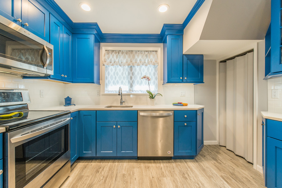 This is an example of a classic l-shaped enclosed kitchen in New York with a submerged sink, shaker cabinets, blue cabinets, white splashback, metro tiled splashback, stainless steel appliances, light hardwood flooring, beige floors and beige worktops.