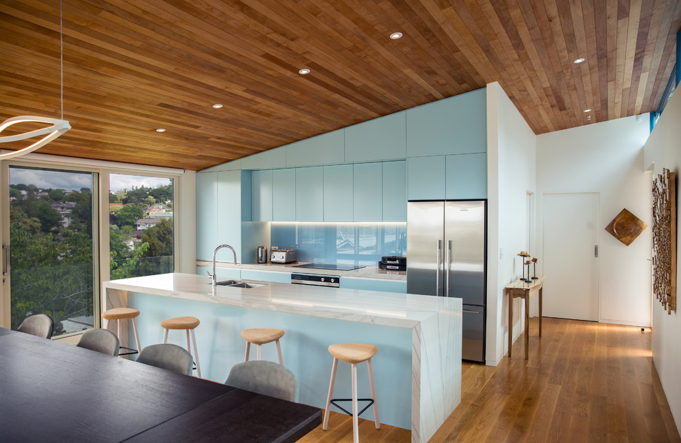 Example of a trendy galley medium tone wood floor and brown floor open concept kitchen design in Auckland with an undermount sink, flat-panel cabinets, blue cabinets, blue backsplash, glass sheet backsplash, stainless steel appliances, an island and white countertops