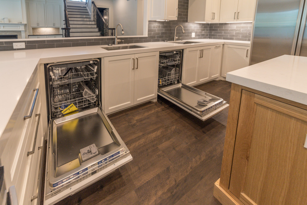 Photo of a large traditional u-shaped open plan kitchen in Houston with a submerged sink, shaker cabinets, white cabinets, quartz worktops, grey splashback, metro tiled splashback, stainless steel appliances, laminate floors, an island, brown floors and white worktops.