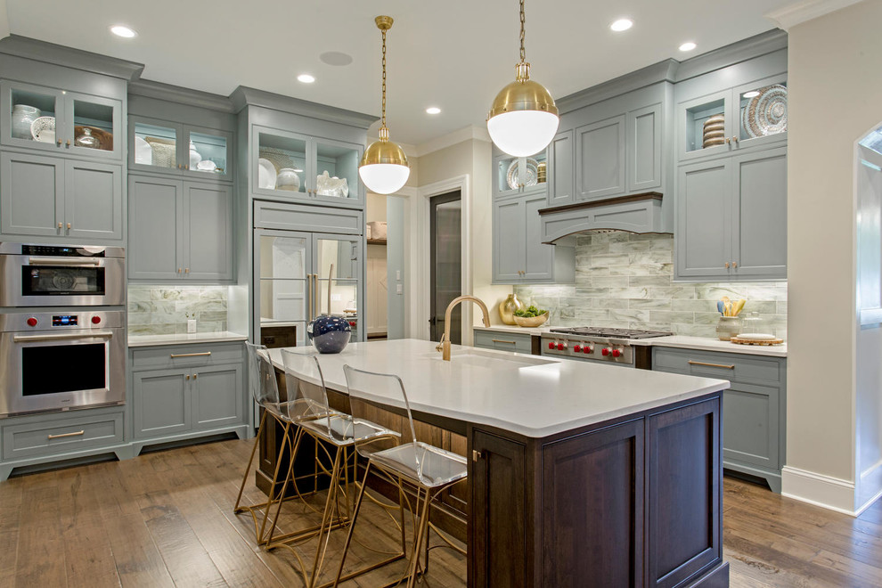 Klassisk inredning av ett kök, med en undermonterad diskho, blå skåp, grått stänkskydd, rostfria vitvaror, mörkt trägolv, en köksö, luckor med profilerade fronter och bänkskiva i kvarts
