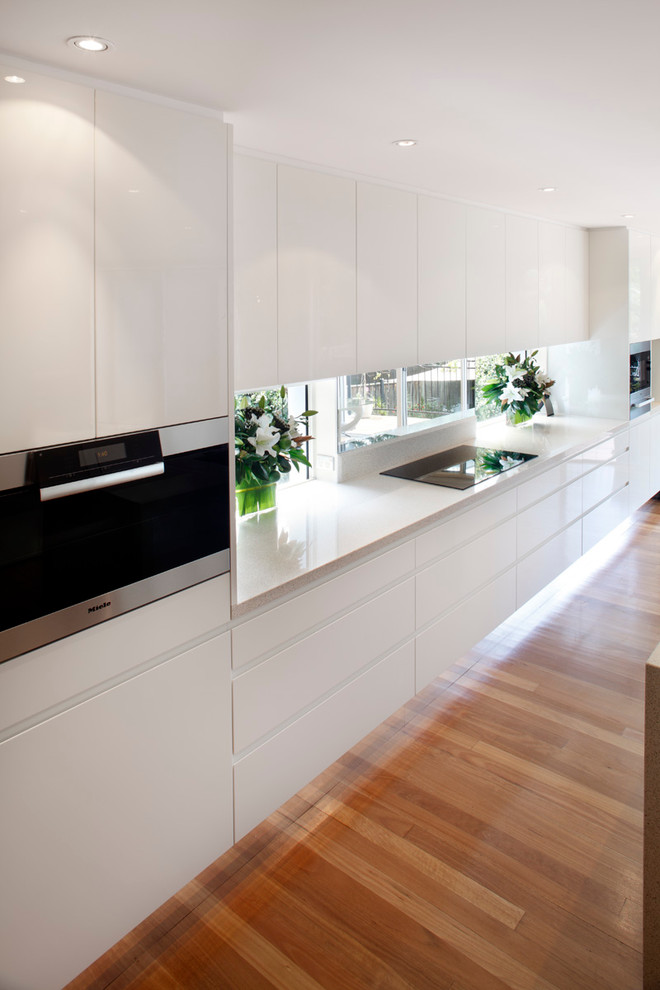 Inspiration for a small contemporary galley open plan kitchen in Sydney with a submerged sink, flat-panel cabinets, white cabinets, quartz worktops, white splashback, glass sheet splashback, stainless steel appliances, medium hardwood flooring and an island.