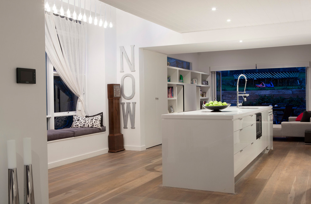 Example of a mid-sized trendy galley light wood floor and brown floor open concept kitchen design in Sydney with an undermount sink, flat-panel cabinets, white cabinets, quartz countertops, white backsplash, glass sheet backsplash, stainless steel appliances, an island and white countertops