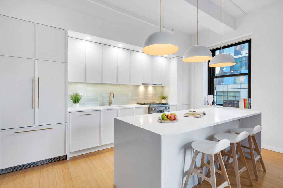 This is an example of a contemporary galley kitchen in New York with a submerged sink, white cabinets, engineered stone countertops, green splashback, integrated appliances, light hardwood flooring, white worktops, recessed-panel cabinets, an island and beige floors.