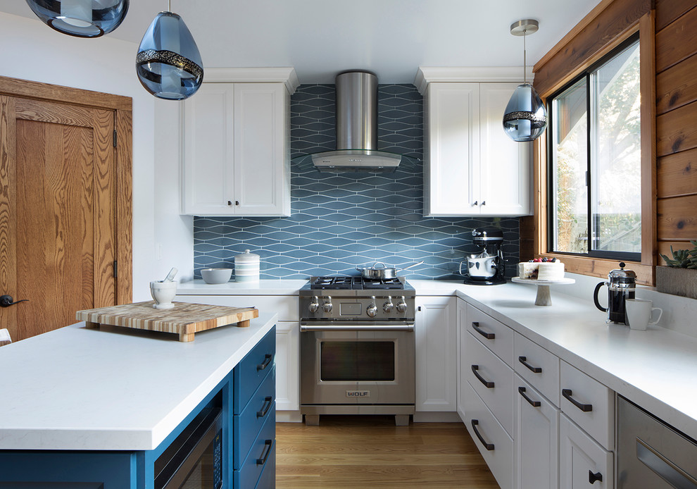 На фото: отдельная, угловая кухня в морском стиле с фасадами с утопленной филенкой, белыми фасадами, синим фартуком, техникой из нержавеющей стали, светлым паркетным полом, островом и окном