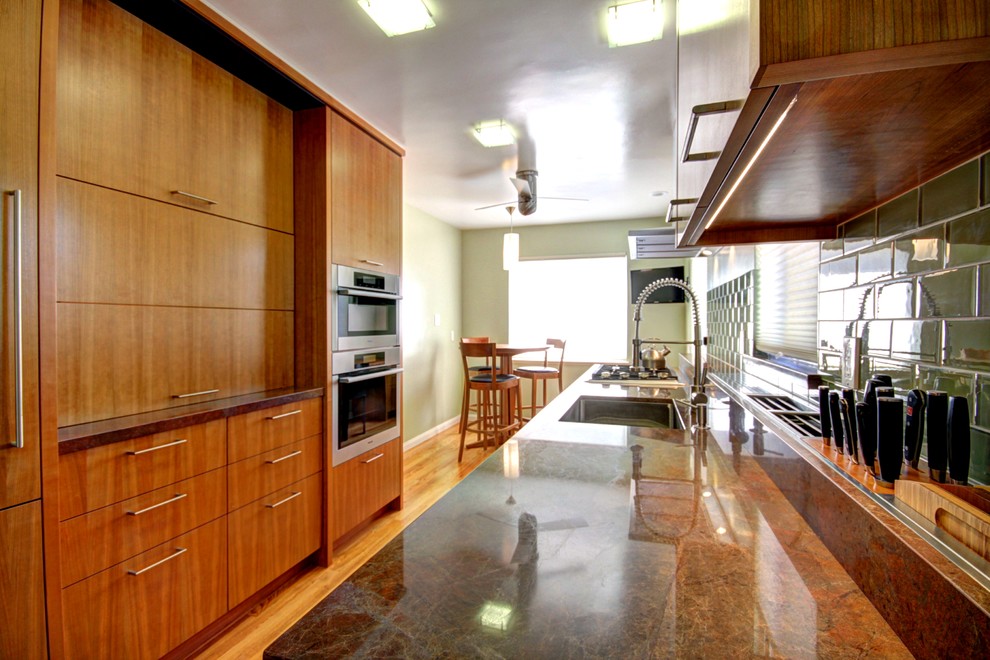 Example of a small trendy galley medium tone wood floor enclosed kitchen design in San Francisco with a single-bowl sink, flat-panel cabinets, medium tone wood cabinets, green backsplash, ceramic backsplash, stainless steel appliances and solid surface countertops