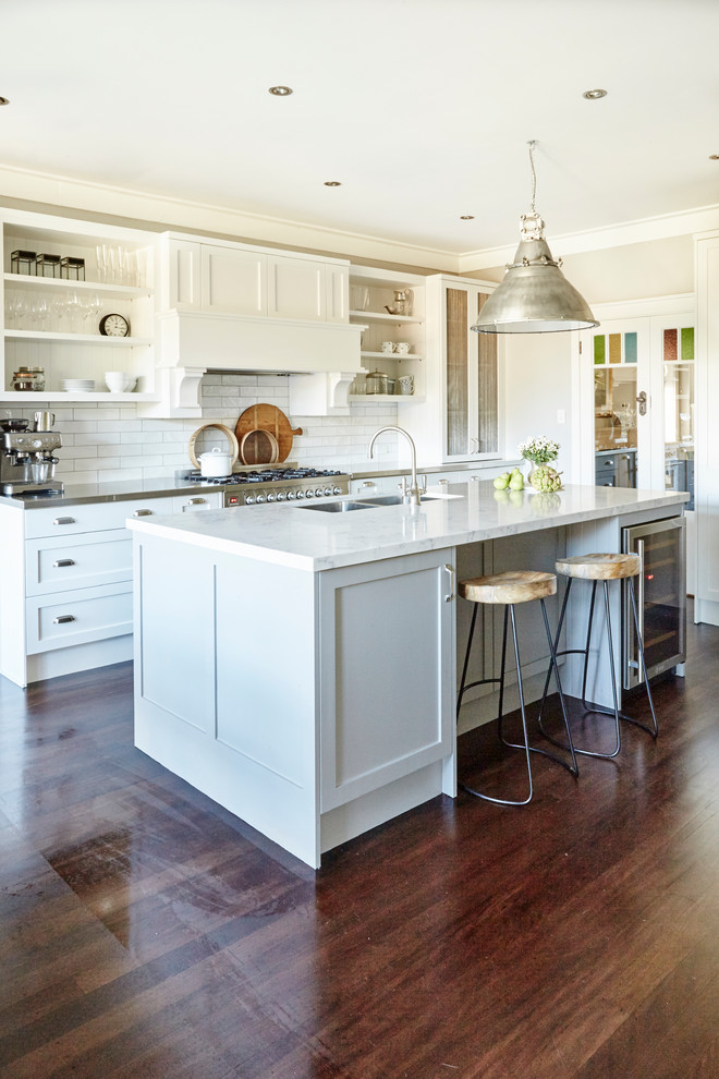 Large traditional l-shaped kitchen/diner in Sydney with a double-bowl sink, shaker cabinets, white cabinets, engineered stone countertops, white splashback, ceramic splashback, stainless steel appliances, medium hardwood flooring and an island.