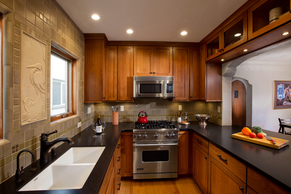 Inredning av ett amerikanskt litet svart svart kök, med en dubbel diskho, skåp i shakerstil, skåp i mellenmörkt trä, granitbänkskiva, grönt stänkskydd, stänkskydd i keramik, rostfria vitvaror, mellanmörkt trägolv, en halv köksö och brunt golv