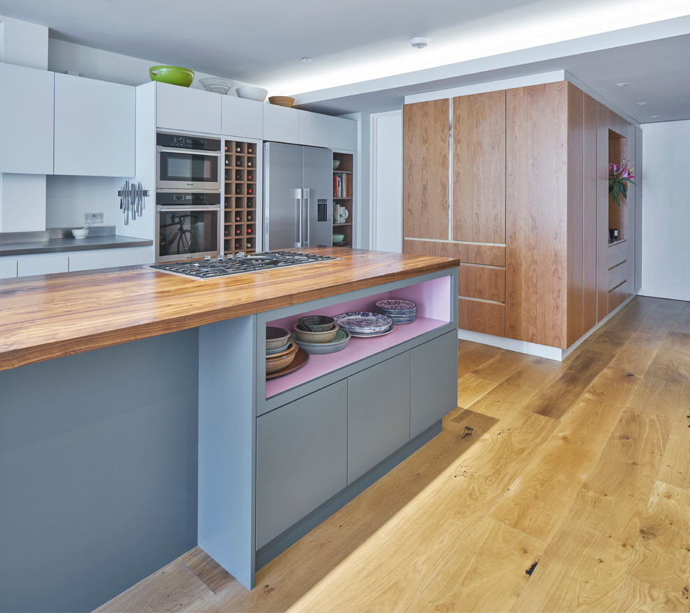 Example of a large trendy light wood floor open concept kitchen design in London with flat-panel cabinets, blue cabinets, stainless steel countertops, paneled appliances and an island