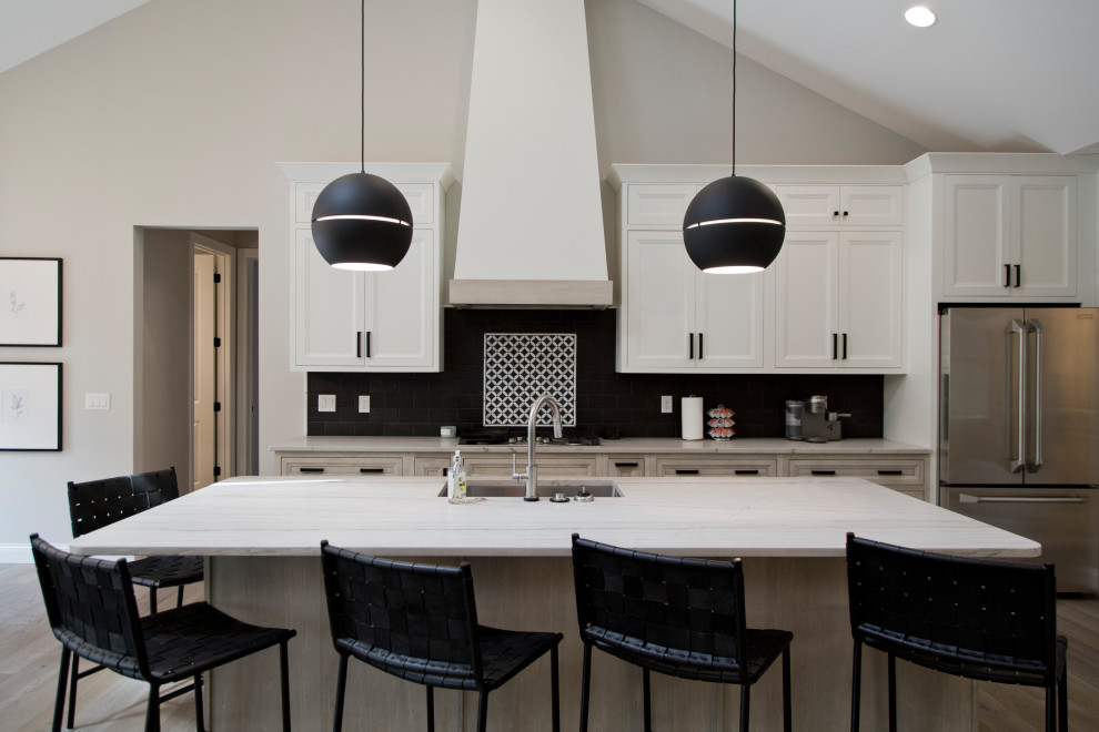 Inspiration for a large classic l-shaped open plan kitchen in St Louis with a submerged sink, recessed-panel cabinets, white cabinets, engineered stone countertops, black splashback, ceramic splashback, stainless steel appliances, light hardwood flooring, an island, beige floors and white worktops.
