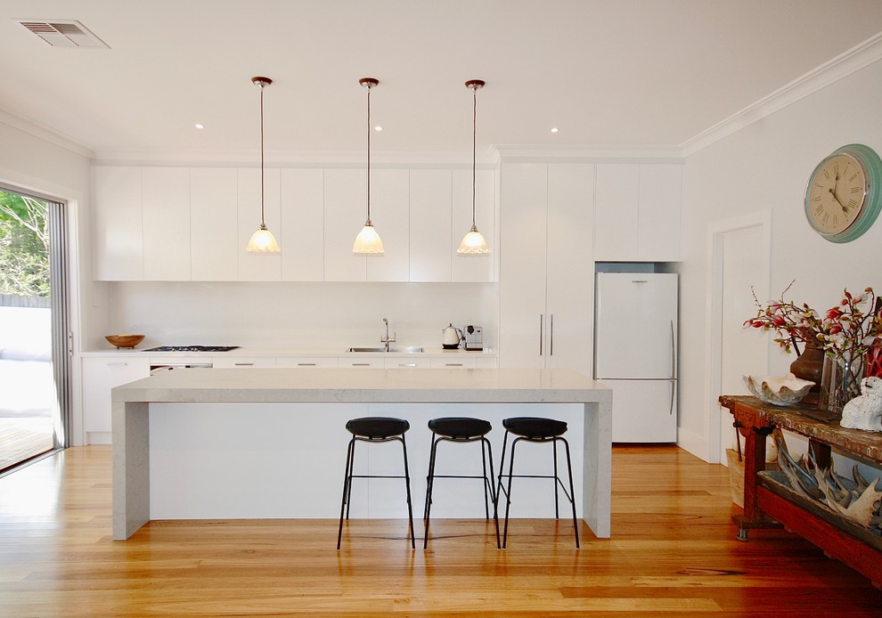 Large modern single-wall kitchen/diner in Sydney with a built-in sink, flat-panel cabinets, white cabinets, engineered stone countertops, white splashback, stone slab splashback, white appliances, medium hardwood flooring, an island and brown floors.