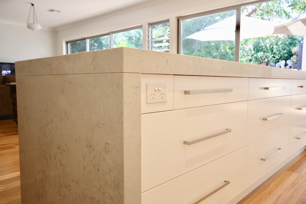 This is an example of a large modern single-wall kitchen/diner in Sydney with a built-in sink, flat-panel cabinets, white cabinets, engineered stone countertops, white splashback, stone slab splashback, white appliances, medium hardwood flooring, an island and brown floors.