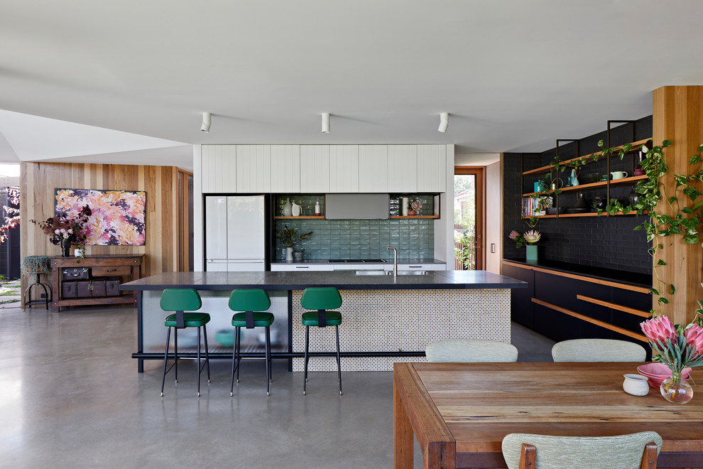 На фото: прямая кухня-гостиная в скандинавском стиле с накладной мойкой, плоскими фасадами, белыми фасадами, зеленым фартуком, белой техникой, бетонным полом, островом, серым полом и черной столешницей с