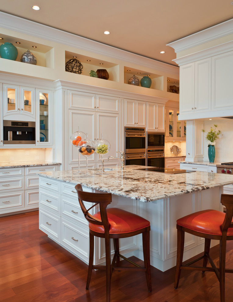 Example of a large classic l-shaped medium tone wood floor and beige floor enclosed kitchen design in Miami with an undermount sink, recessed-panel cabinets, white cabinets, granite countertops, white backsplash, stone tile backsplash, paneled appliances and an island