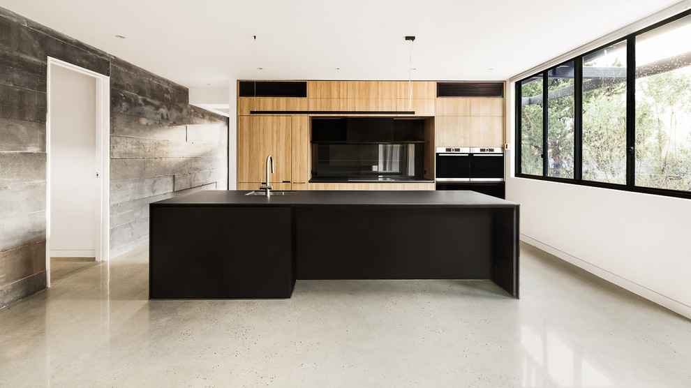 Design ideas for a contemporary kitchen in Melbourne with a submerged sink, glass sheet splashback, black appliances, concrete flooring, flat-panel cabinets, light wood cabinets, black splashback and an island.