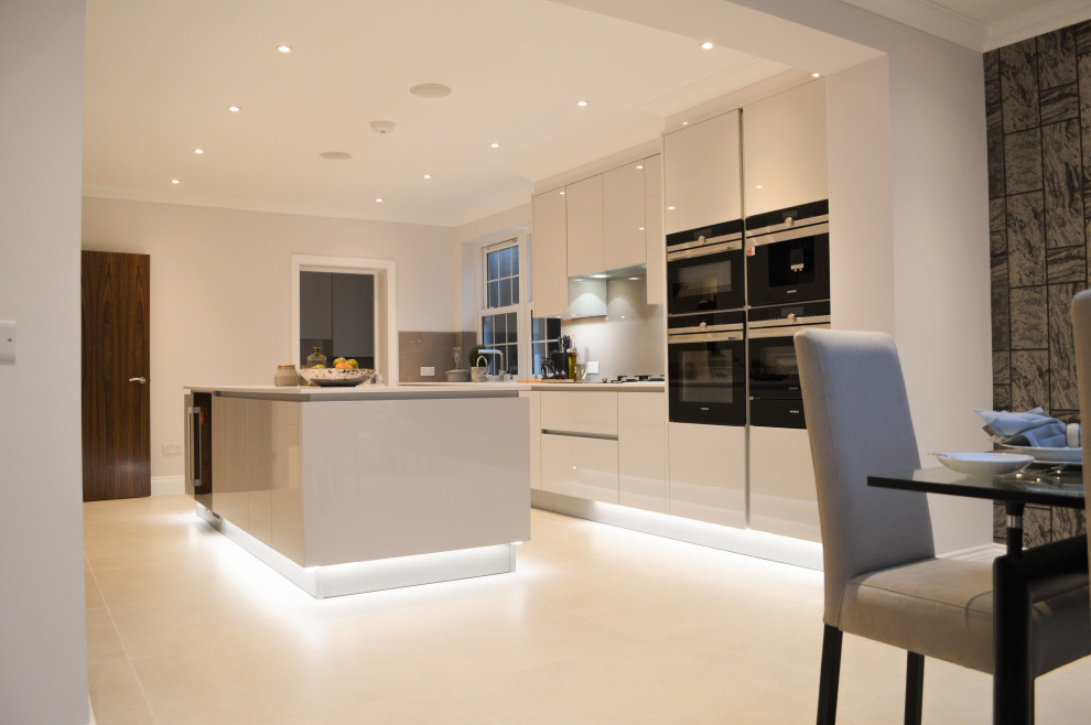 Photo of a large contemporary galley kitchen/diner in London with a built-in sink, flat-panel cabinets, white cabinets, white splashback, porcelain splashback, black appliances, an island and white worktops.