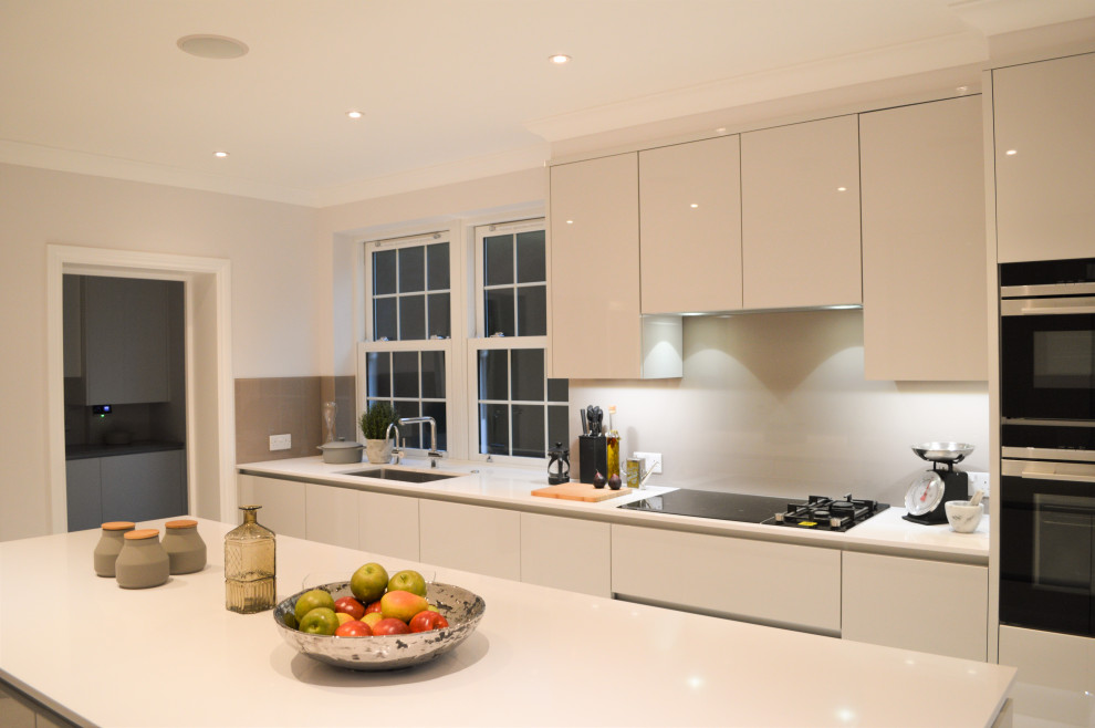 Design ideas for a large contemporary galley kitchen/diner in London with a built-in sink, flat-panel cabinets, white cabinets, white splashback, porcelain splashback, black appliances, an island and white worktops.