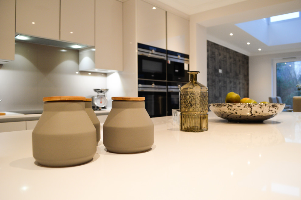Large contemporary galley kitchen/diner in London with a built-in sink, flat-panel cabinets, white cabinets, white splashback, porcelain splashback, black appliances, an island and white worktops.