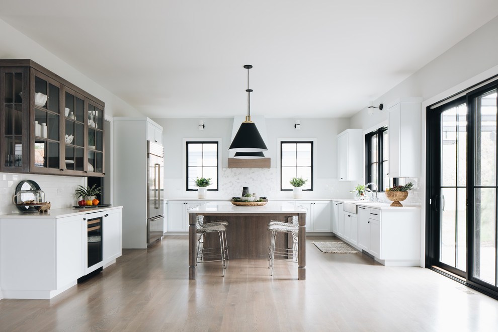 Inspiration for a traditional u-shaped kitchen in Chicago with a belfast sink, shaker cabinets, white splashback, stainless steel appliances, medium hardwood flooring, an island, brown floors and white worktops.