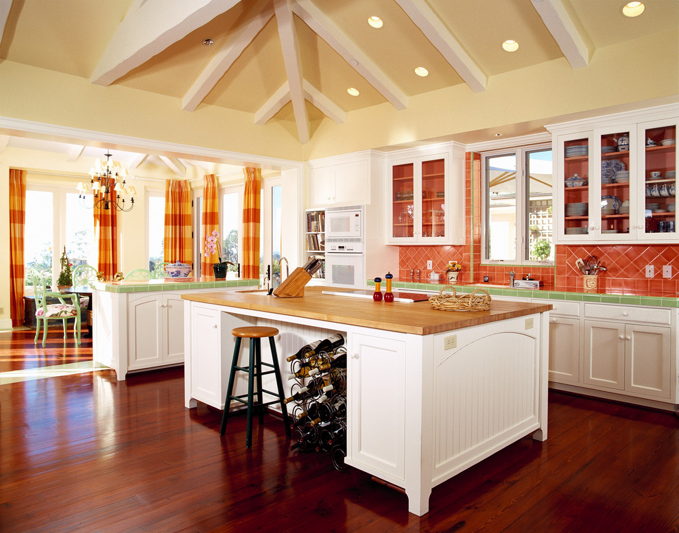 Modelo de cocina clásica abierta con armarios tipo vitrina, puertas de armario blancas, encimera de azulejos, salpicadero naranja, salpicadero de azulejos de cerámica y electrodomésticos blancos