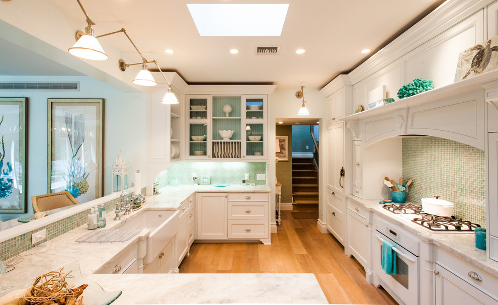 Design ideas for a coastal u-shaped kitchen in Miami with a belfast sink, recessed-panel cabinets, white cabinets, green splashback, mosaic tiled splashback, white appliances, medium hardwood flooring and a breakfast bar.