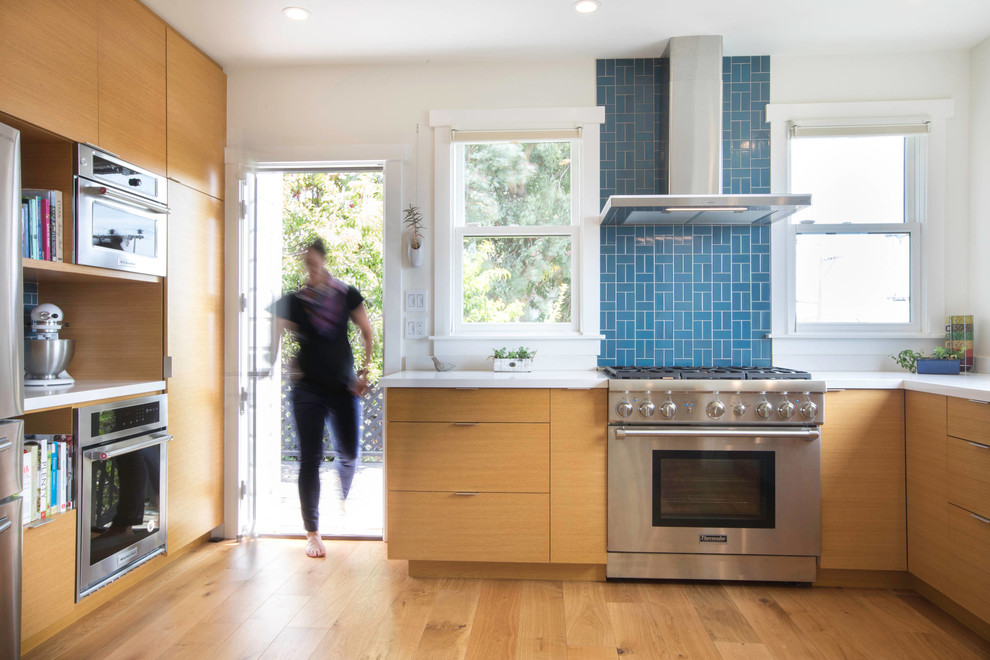 Cette image montre une cuisine design en bois clair avec une crédence bleue, un plan de travail blanc, un placard à porte plane, un électroménager en acier inoxydable, parquet clair et aucun îlot.