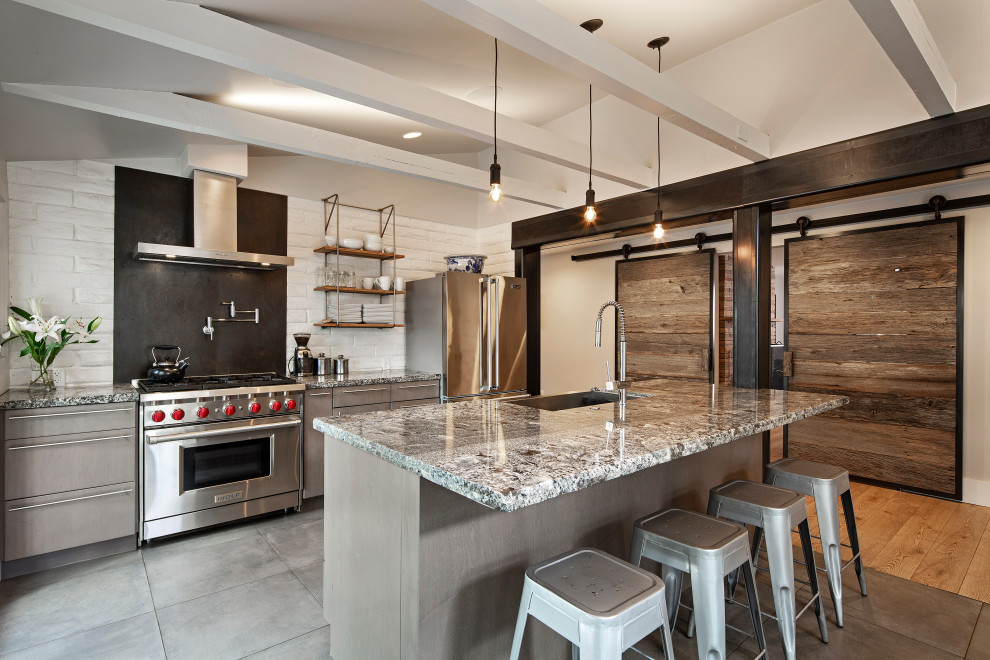 Bild på ett rustikt grå grått kök och matrum, med en undermonterad diskho, släta luckor, grå skåp, granitbänkskiva, vitt stänkskydd, stänkskydd i tegel, rostfria vitvaror, cementgolv, en köksö och grått golv