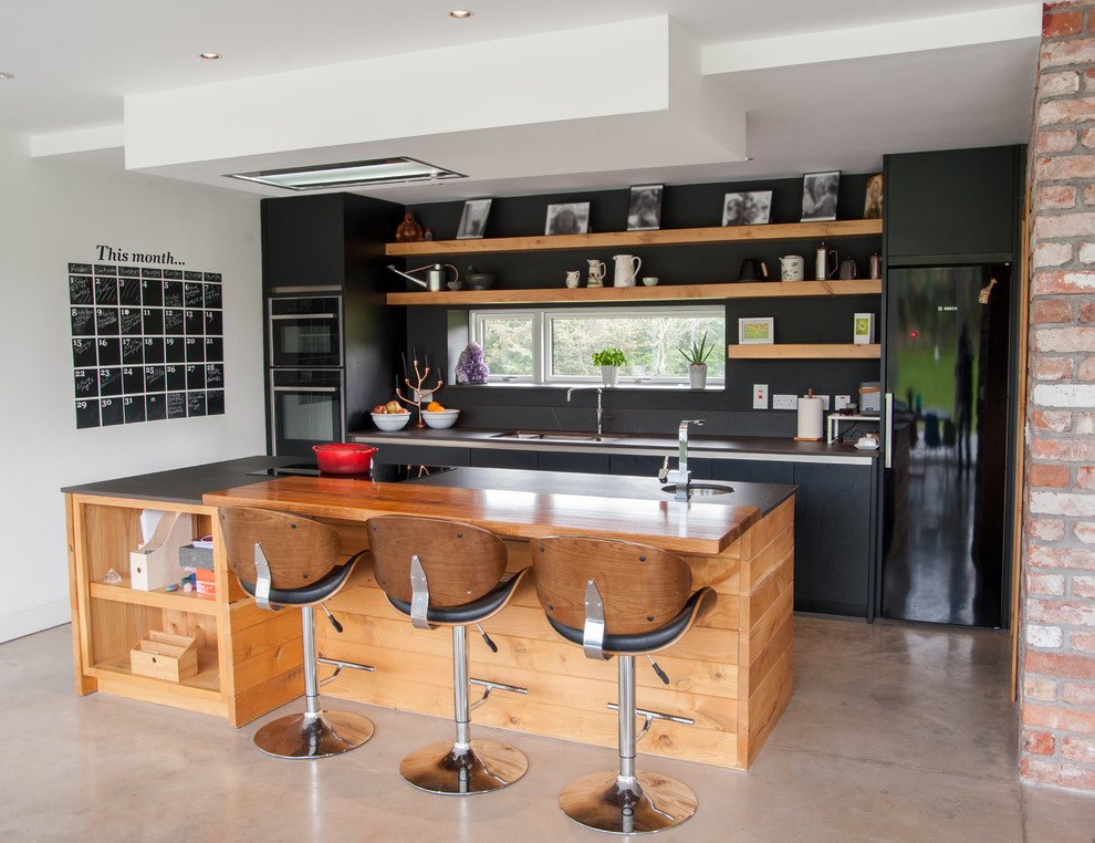 Inspiration for a large industrial galley open plan kitchen in Cork with flat-panel cabinets, black cabinets, black appliances, concrete flooring, an island, grey floors, a double-bowl sink, quartz worktops, brown splashback and brown worktops.