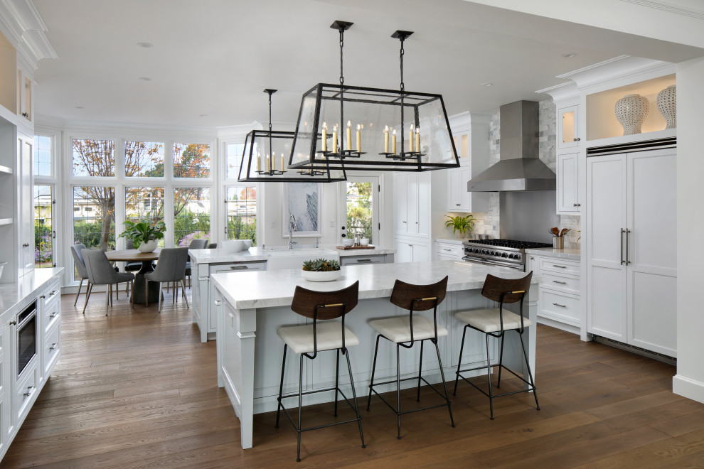 Elegant u-shaped medium tone wood floor and brown floor eat-in kitchen photo in San Francisco with a farmhouse sink, recessed-panel cabinets, white cabinets, gray backsplash, subway tile backsplash, paneled appliances, two islands and gray countertops