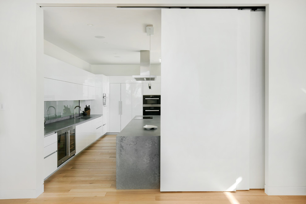 Modern inredning av ett stort grå grått kök, med en undermonterad diskho, släta luckor, vita skåp, bänkskiva i kvarts, glaspanel som stänkskydd, integrerade vitvaror, ljust trägolv, en köksö och brunt golv