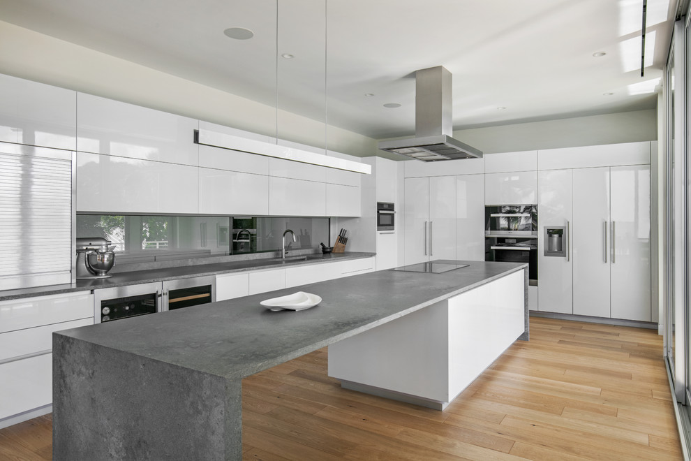 Modern inredning av ett stort grå grått kök, med en undermonterad diskho, släta luckor, vita skåp, bänkskiva i kvarts, glaspanel som stänkskydd, integrerade vitvaror, ljust trägolv, en köksö, brunt golv och grått stänkskydd