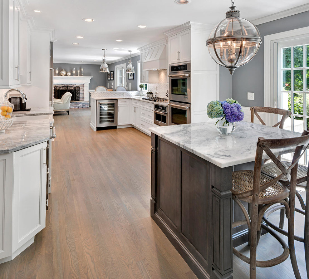 Example of a large transitional l-shaped light wood floor enclosed kitchen design in New York with a farmhouse sink, recessed-panel cabinets, gray cabinets, marble countertops, white backsplash, stone tile backsplash, stainless steel appliances and an island