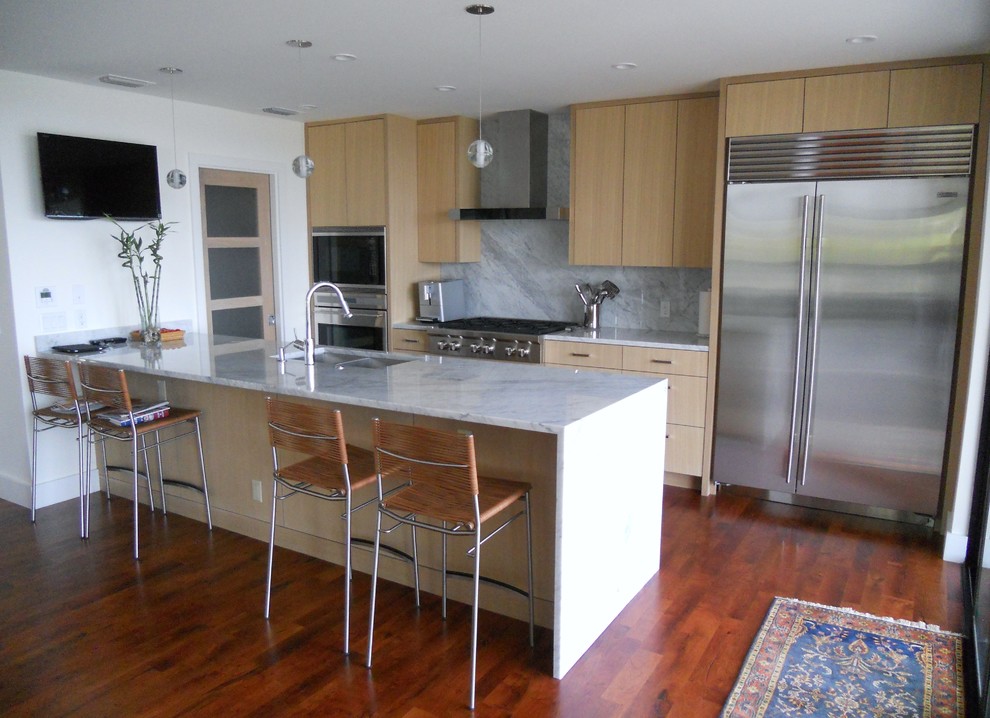 Modelo de cocina comedor actual con armarios con paneles lisos, puertas de armario de madera clara, salpicadero amarillo, electrodomésticos de acero inoxidable, suelo de madera oscura y península