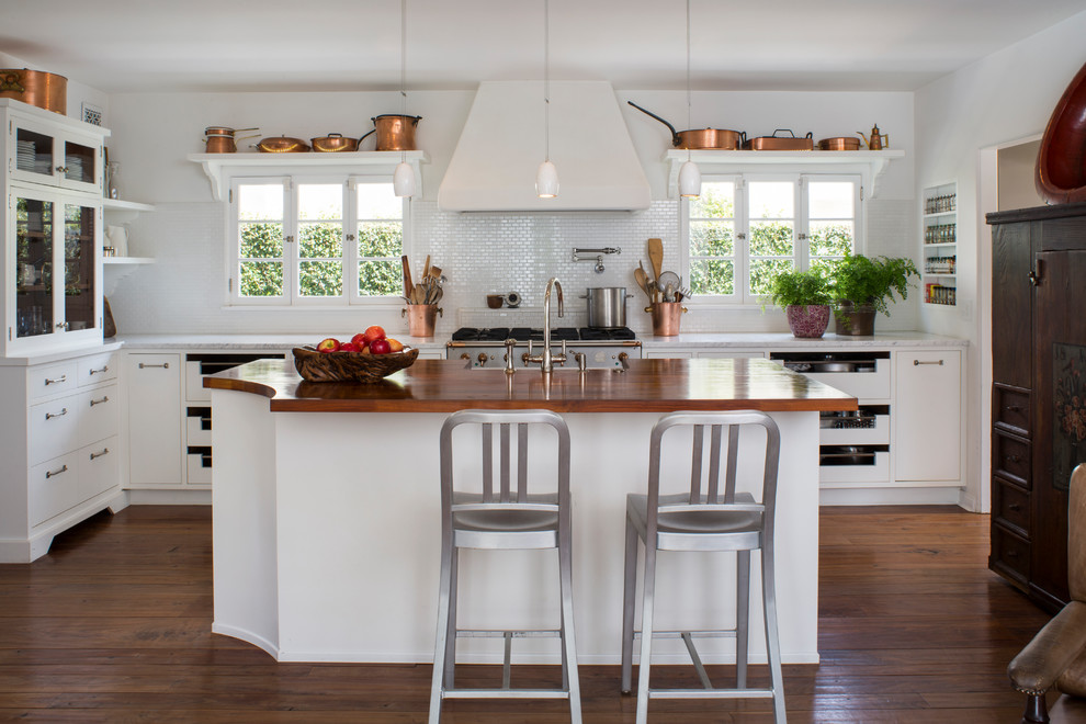 Bild på ett lantligt l-kök, med en rustik diskho, släta luckor, vita skåp, vitt stänkskydd, stänkskydd i mosaik, rostfria vitvaror, mörkt trägolv, en köksö och brunt golv