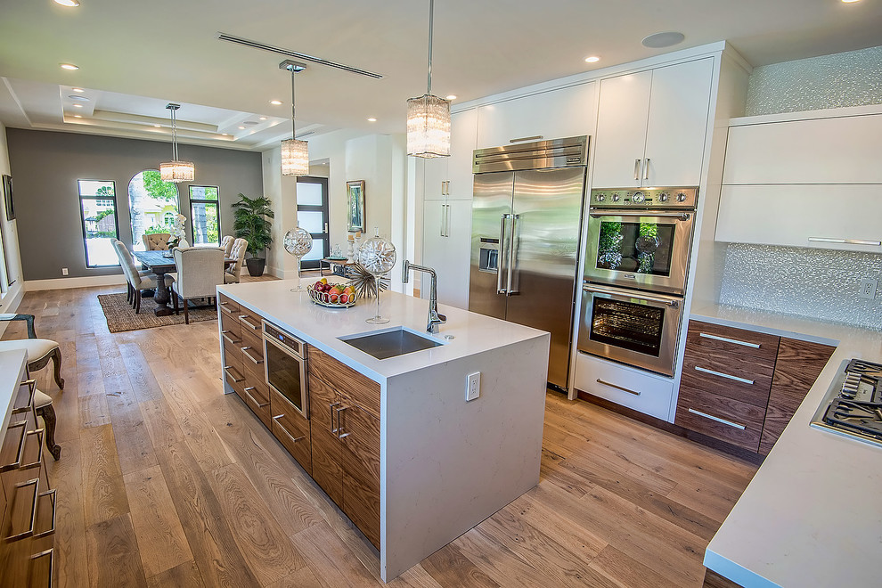 Large modern u-shaped kitchen/diner in Los Angeles with a submerged sink, flat-panel cabinets, white cabinets, composite countertops, white splashback, stainless steel appliances, medium hardwood flooring, an island and brown floors.