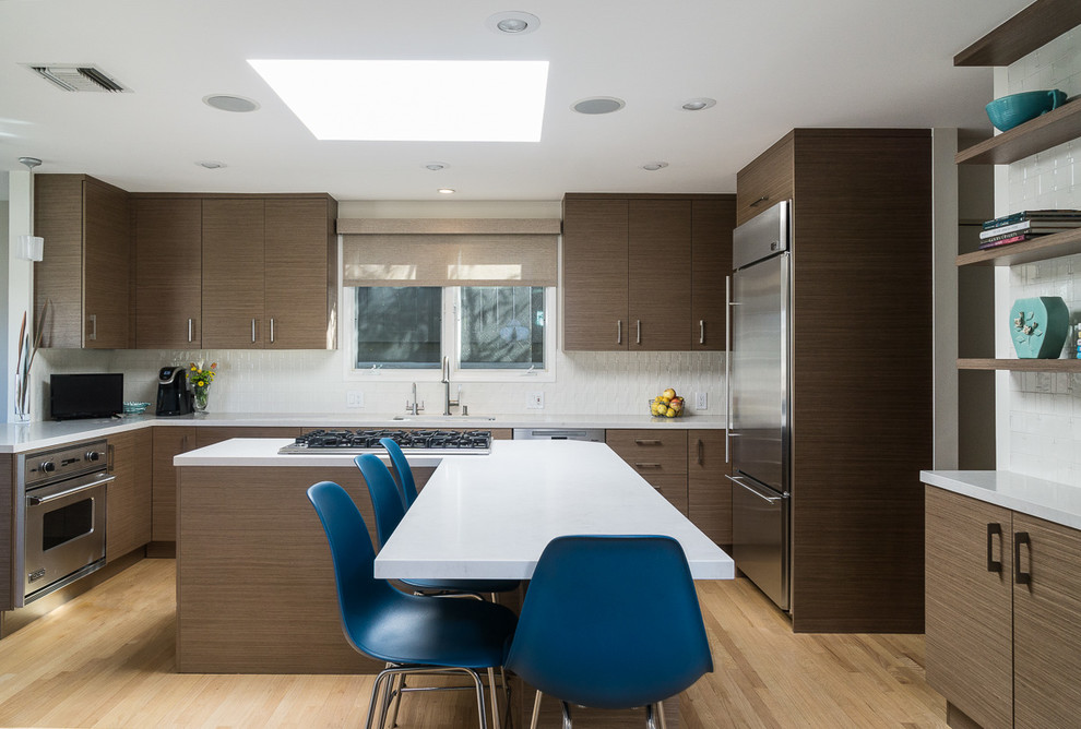 Moderne Küche in L-Form mit flächenbündigen Schrankfronten, dunklen Holzschränken, Mineralwerkstoff-Arbeitsplatte, Küchenrückwand in Weiß, Küchengeräten aus Edelstahl, hellem Holzboden, Kücheninsel und braunem Boden in Los Angeles