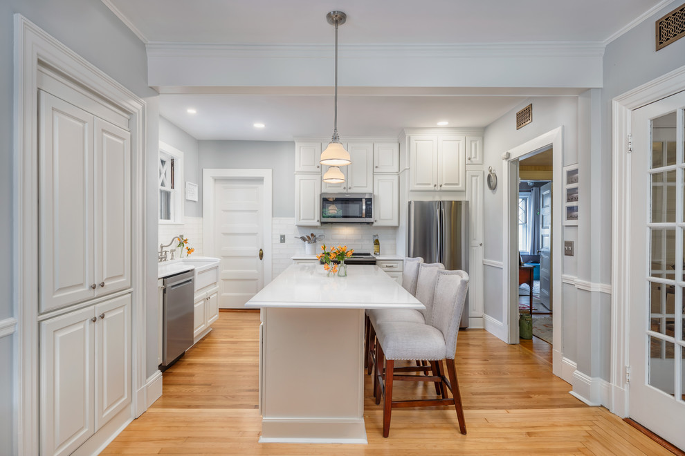 Example of a mid-sized classic l-shaped medium tone wood floor and beige floor eat-in kitchen design in Boston with a farmhouse sink, raised-panel cabinets, white cabinets, quartzite countertops, white backsplash, subway tile backsplash, stainless steel appliances, an island and white countertops