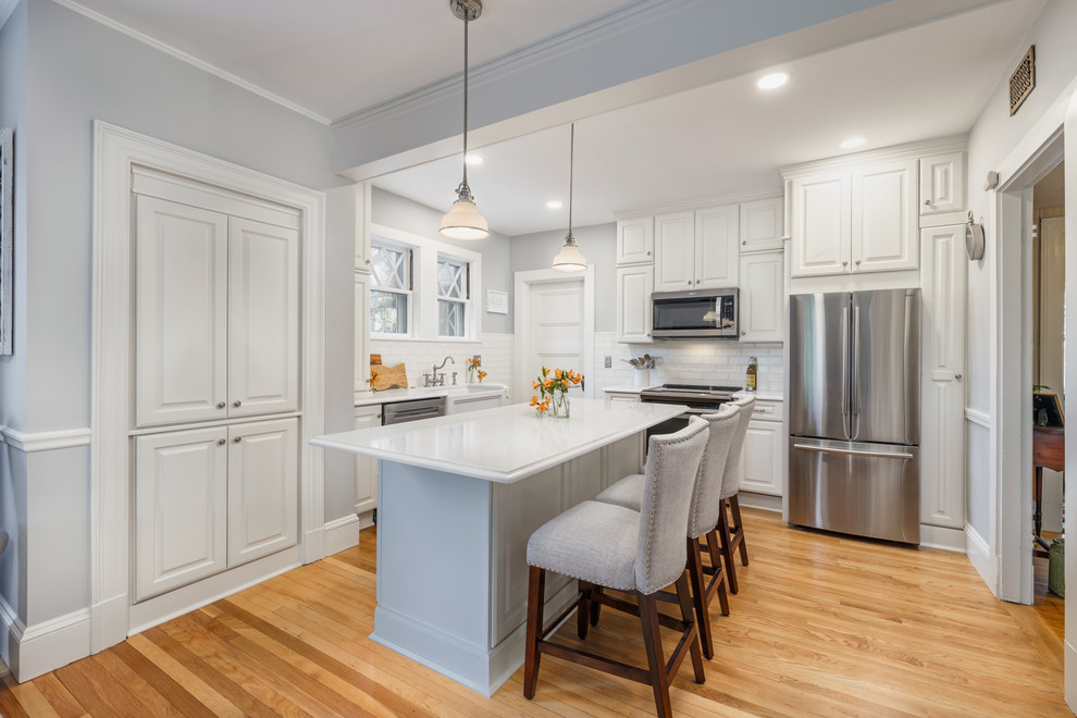 Inspiration for a mid-sized timeless l-shaped medium tone wood floor and beige floor eat-in kitchen remodel in Boston with a farmhouse sink, raised-panel cabinets, white cabinets, quartzite countertops, white backsplash, subway tile backsplash, stainless steel appliances, an island and white countertops